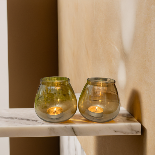 Two glass candle holders with lit tealight candles on a marble surface against a neutral wall.