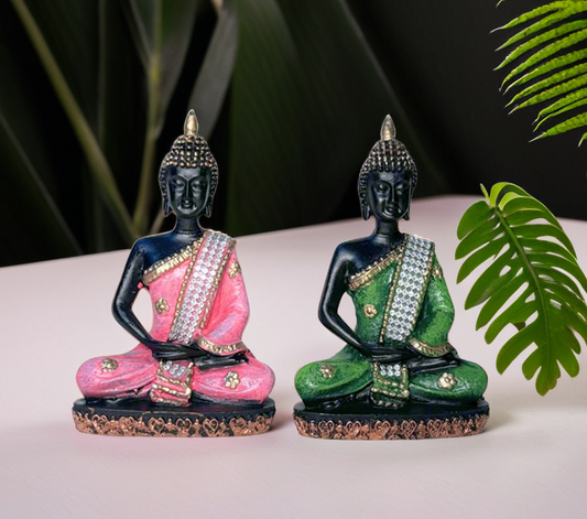 Two decorative Buddha statues with pink and green outfits on a white surface with leaves in the background.