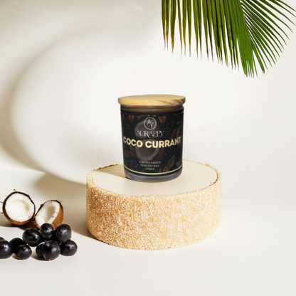 Candle labeled 'Coco Currant' on a decorative stand with coconut halves and black fruits, palm leaf in the background.
