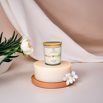 A scented glass candle jar with a wooden lid, labeled 'valley of flowers', surrounded by decorative flowers and petals on a beige background.