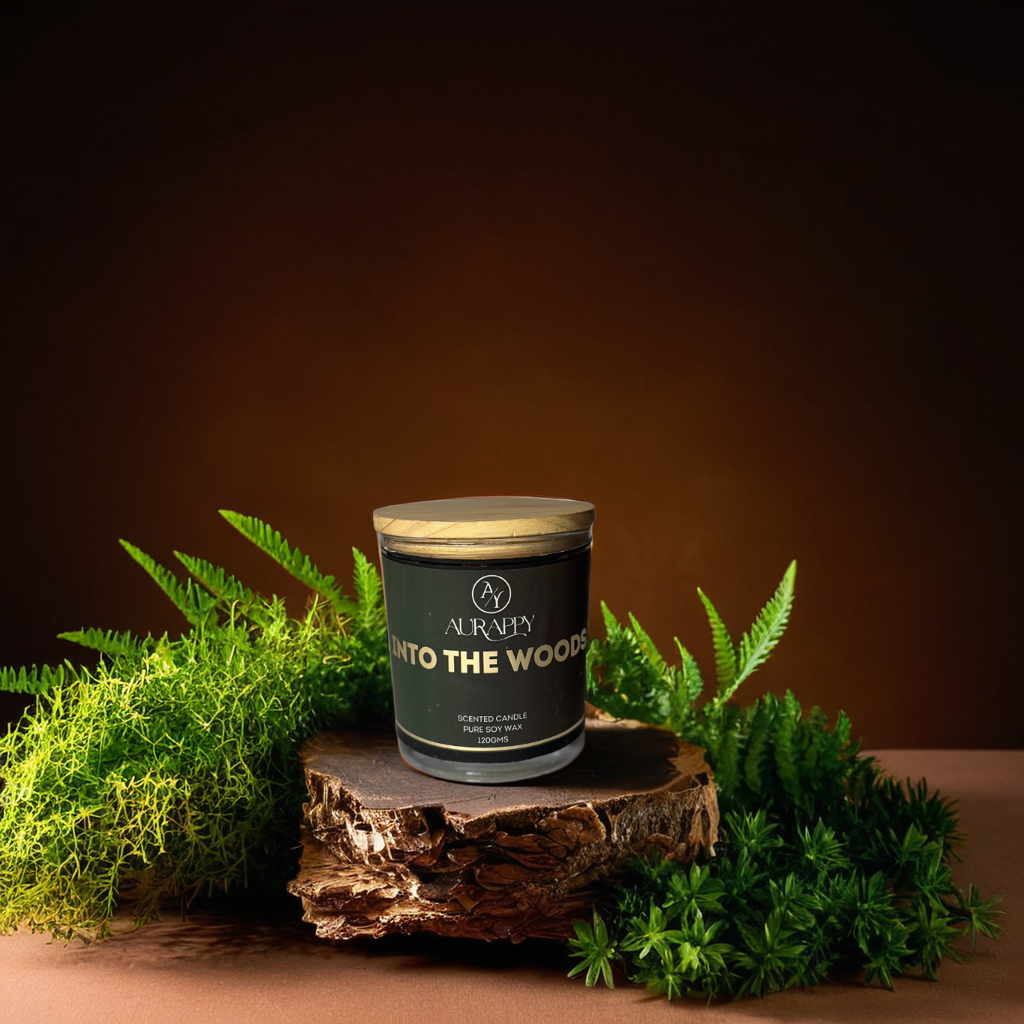 A scented glass candle jar with a wooden lid, labeled 'into the woods', surrounded by decorative grass and petals on a brown background.